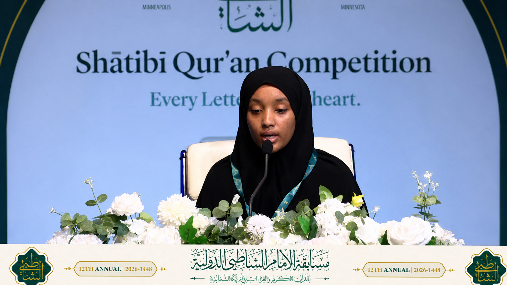 A student reciting Qur'an into a microphone on stage at the Shatibi Qur'an Competition